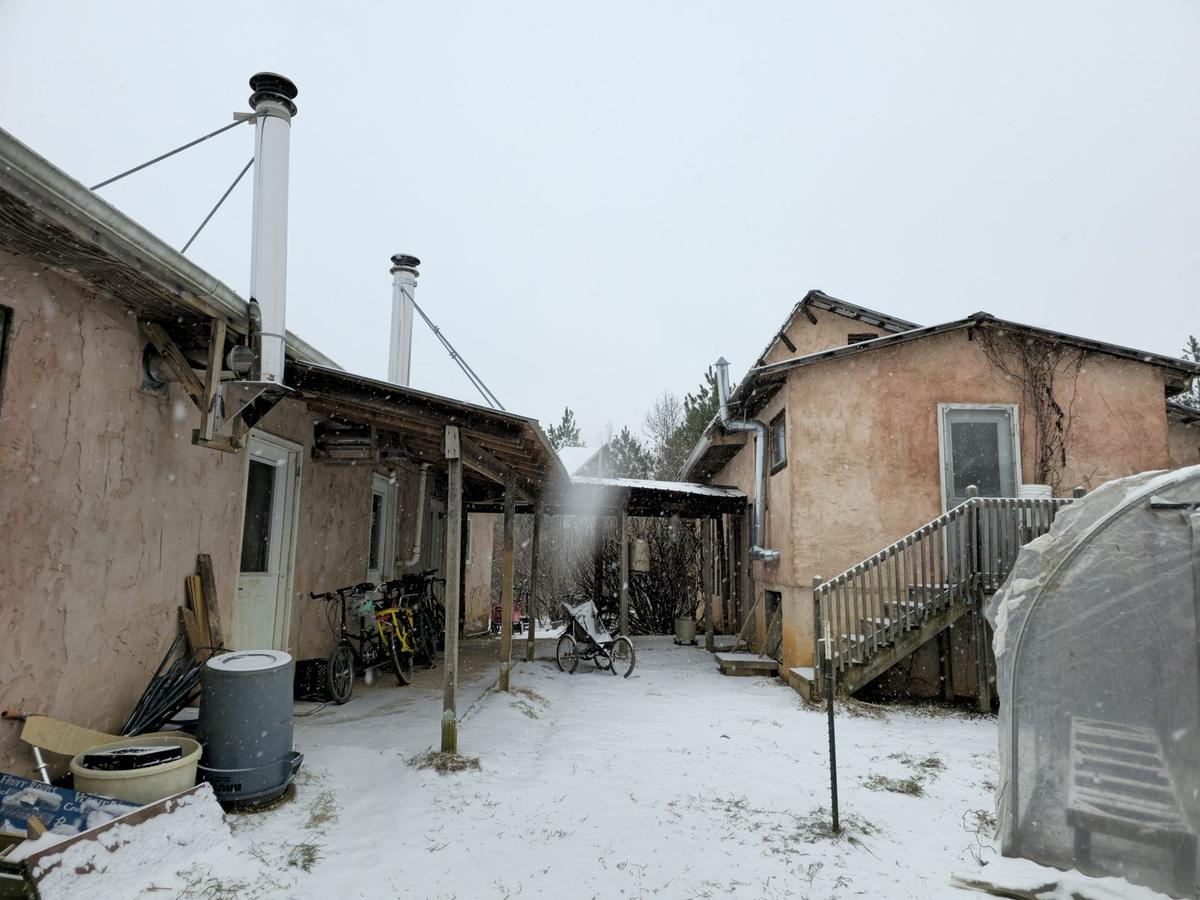 Two plastered houses around a snowy courtyard at Living Energy Farm, with stovepipes and a greenhouse.